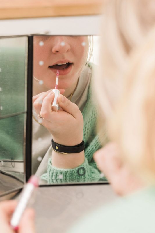 someone applying custom, hydrating lip gloss in front of a mirror to try plumping hyaluronic acid lip gloss formula made with plant-based ingredients